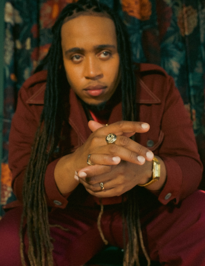 Portrait of drummer and singer Jamison Ross, African American man, approx 30, long hair, maroon outfit.